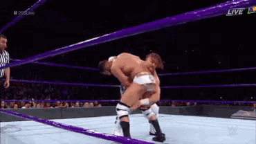 two men are wrestling in a wrestling ring with purple ropes and a referee watching .
