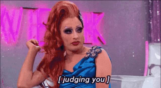 a drag queen is sitting in front of a pink sign and says i judging you .