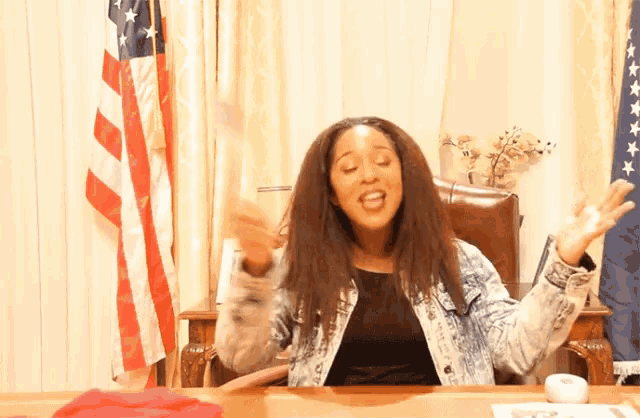 a woman in a denim jacket is sitting at a desk with an american flag in the background