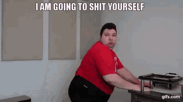 a man in a red shirt and black shorts is standing next to a table and says `` i am going to shit yourself ''