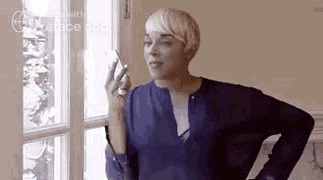 a woman in a blue shirt is standing in front of a window talking on a cell phone .