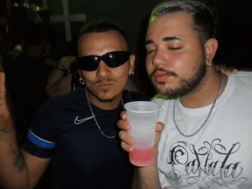 a man holding a cup next to another man wearing sunglasses and a shirt that says cola