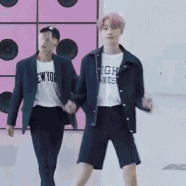 two men are holding hands and dancing in front of a pink wall with speakers .