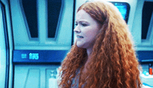 a woman with red curly hair is standing in a room with a computer screen behind her .