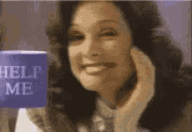 a woman is smiling and talking on a cell phone while holding a help me sign .