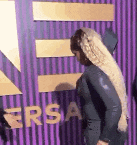 a woman with long blonde hair is standing in front of a purple wall with the word ersa written on it .