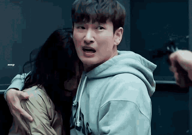 a man in a hoodie is hugging a woman while another man holds a gun in the background .