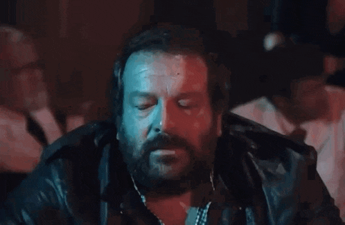 a man with a beard is sitting at a table with his eyes closed and his hands on his face .