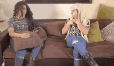 two women are sitting on a couch pillow fighting each other .