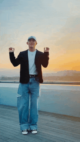 a man in a black jacket and blue jeans is standing on a balcony with his hands in the air