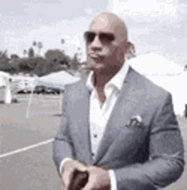 a bald man in a suit and sunglasses is standing in a parking lot holding a cell phone .