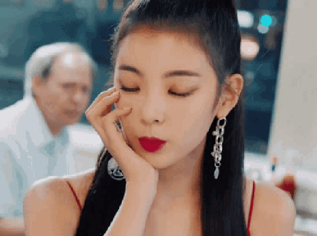a woman wearing red lipstick and earrings looks at the camera with her hand on her face