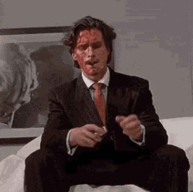 a man in a suit and tie with blood on his face is sitting on a couch smoking a cigarette .
