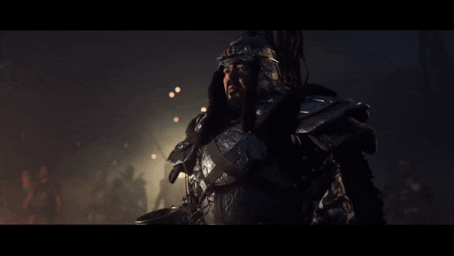 a man in armor stands in a dark room with a group of people in the background