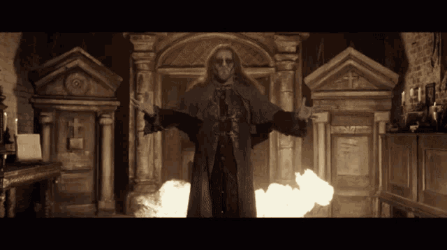 a man with long hair and a beard is standing in a dark room with flames behind him