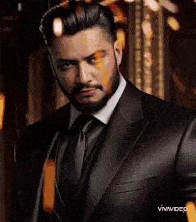 a man with a beard wearing a black suit and tie is looking at the camera .