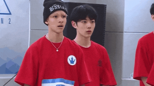 a man wearing a beanie and a red shirt with a crown on it stands next to another man