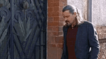 a man with long hair and a beard is standing in front of a brick wall .