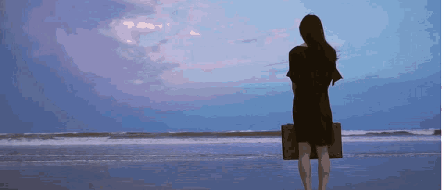 a woman in a black dress is standing on the beach holding a suitcase