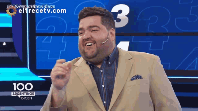 a man in a tan suit is smiling in front of a screen that says 100 argentinas dicen