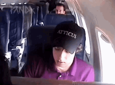 a man wearing a hat that says atticus is sitting on an airplane