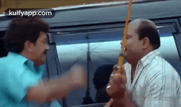 two men are fighting in front of a car and one of them is holding a stick .