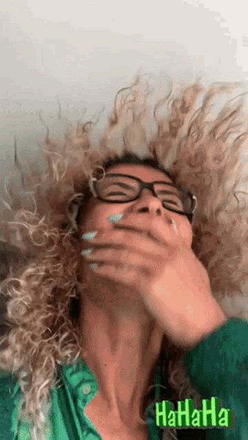 a woman with curly hair and glasses is laughing and covering her mouth with her hand with a green background that says hahaha