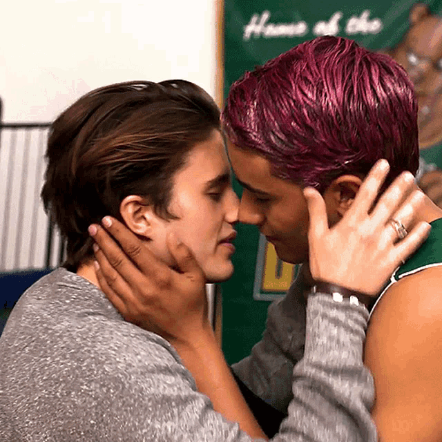 a man with purple hair is kissing another man in front of a banner that says home of the