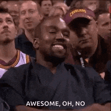 a man is sitting in a crowd of people and smiling . he is wearing a cleveland cavaliers hat .