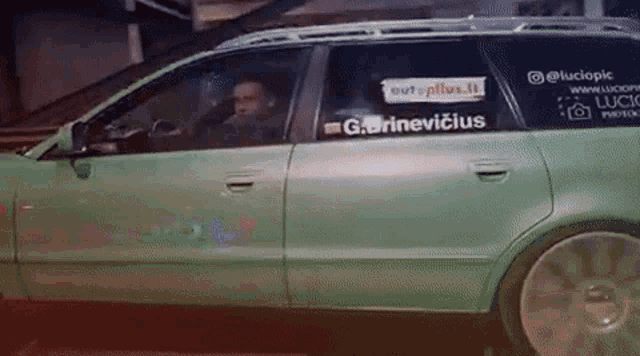 a man is driving a green car with a sticker on the door that says g.grinevicius .