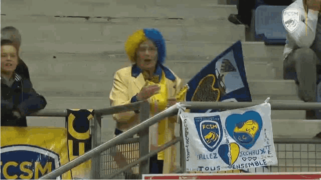 a woman wearing a yellow and blue wig stands in a stadium holding a flag that says fcsm