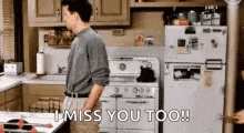 a man is standing in a kitchen with the words `` i miss you too '' written on the fridge .