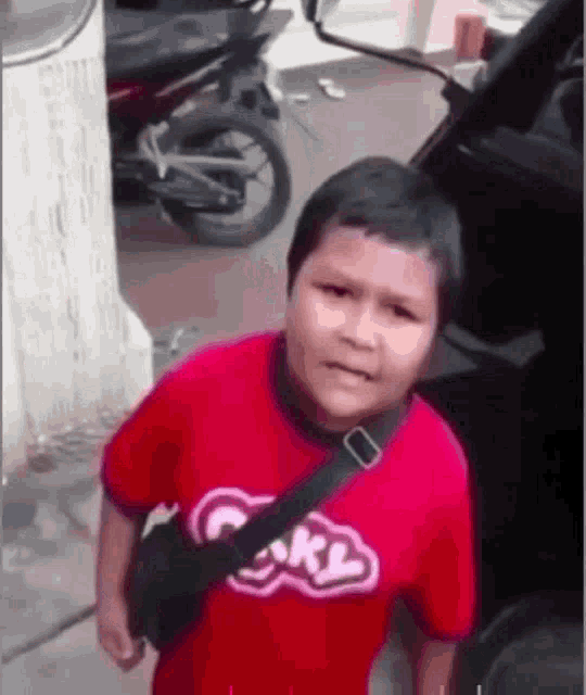 a young boy in a red shirt is standing in front of a car and a motorcycle .