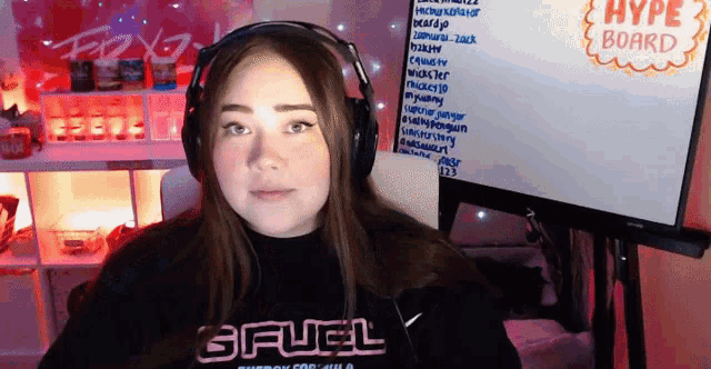 a girl wearing headphones and a sweatshirt that says gfuel stands in front of a hype board