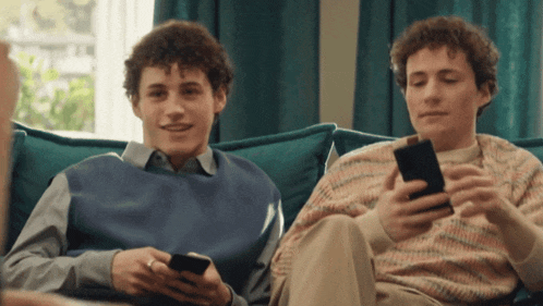 two young men sitting on a couch looking at their cell phones