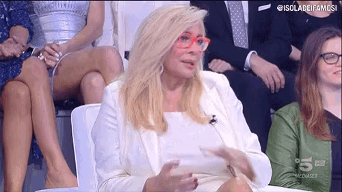 a woman wearing glasses and a white jacket is sitting in front of a group of people .