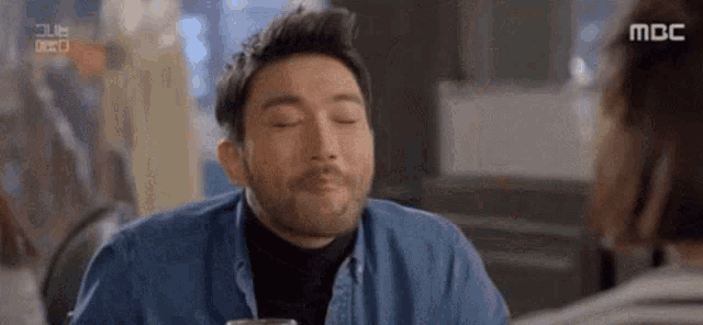 a man with a beard is sitting at a table with his eyes closed and drinking a glass of beer .