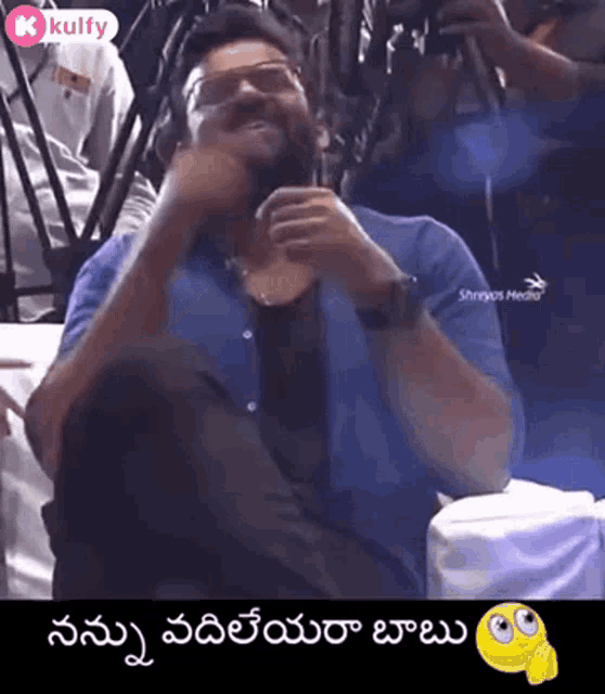 a man wearing glasses and a blue shirt is sitting on a chair and laughing .