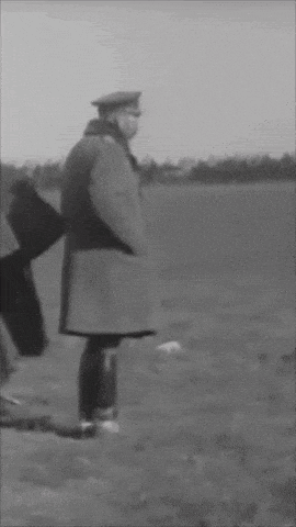 a black and white photo of a man in a trench coat pointing in a field