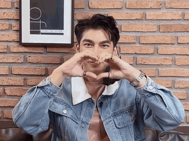 a man in a denim jacket is making a heart shape with his hands in front of a brick wall