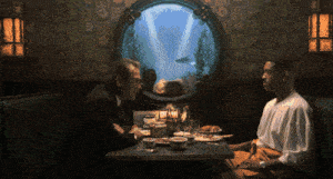 two men sit at a table with a fish tank in the background