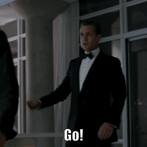 a man in a tuxedo is standing in front of a window and saying go