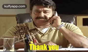 a man with a mustache is sitting at a desk talking on a telephone and saying `` thank you '' .