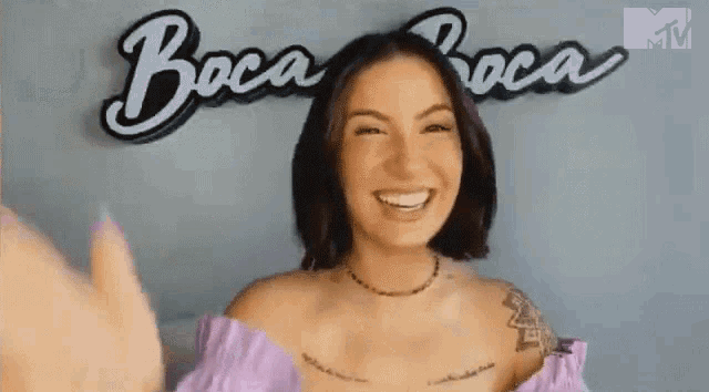 a woman in a purple top is smiling and waving in front of a sign that says boca boca .