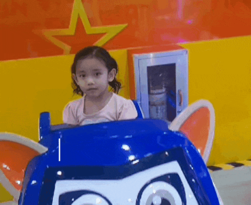 a little girl is sitting in a blue toy car with a star on the wall behind her .