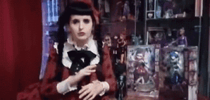 a woman in a red dress and black hat is standing in front of a display case filled with dolls .