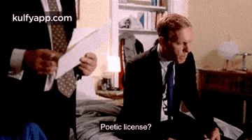a man in a suit and tie is sitting on a bed talking to another man holding a piece of paper .