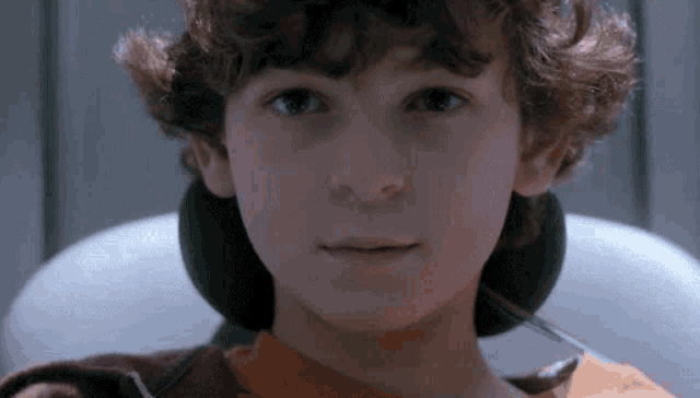 a young boy with curly hair is sitting in a chair and looking at the camera with a thermometer in his mouth .
