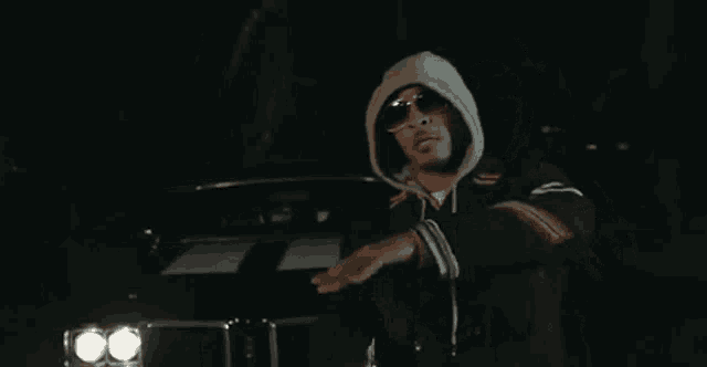 a man in a hooded jacket and sunglasses is standing in front of a black car
