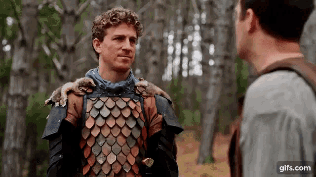 a man in armor holds a squirrel on his shoulder while talking to another man in the woods .